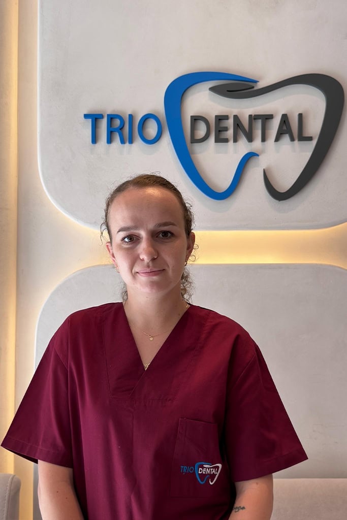 Dentist in maroon scrubs stands in front of Trio Dental logo, showcasing professional dental services.