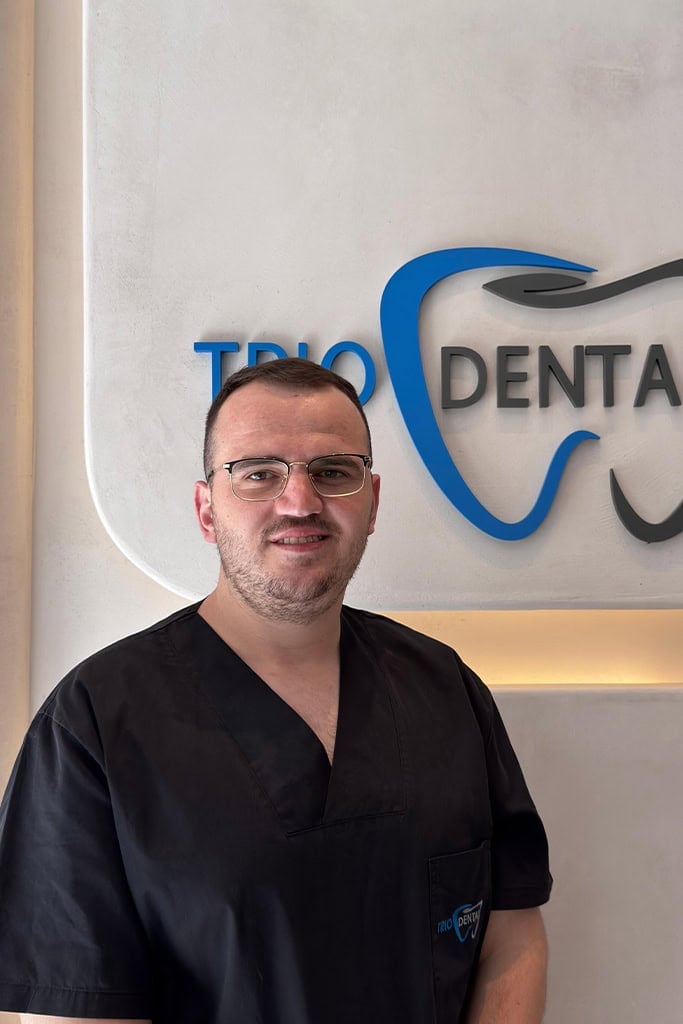 Dentist in black scrubs standing by Trio Dental logo, smiling in a modern clinic setting.