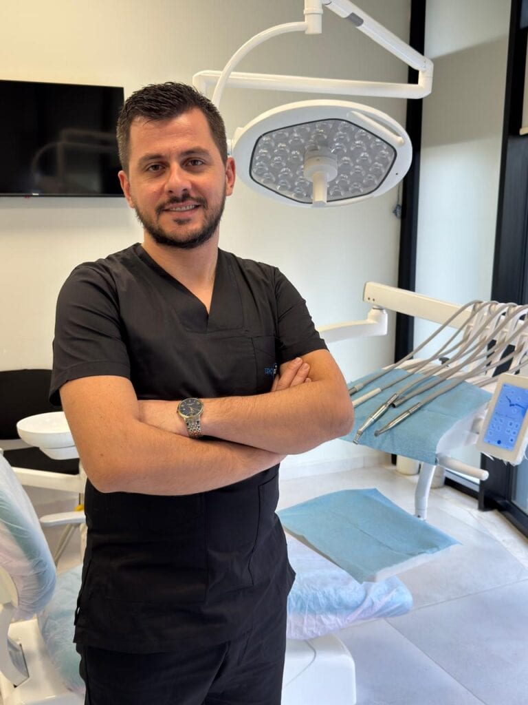 Dentista in camice nero in piedi in un moderno studio dentistico con le braccia conserte e attrezzature dentistiche sullo sfondo.