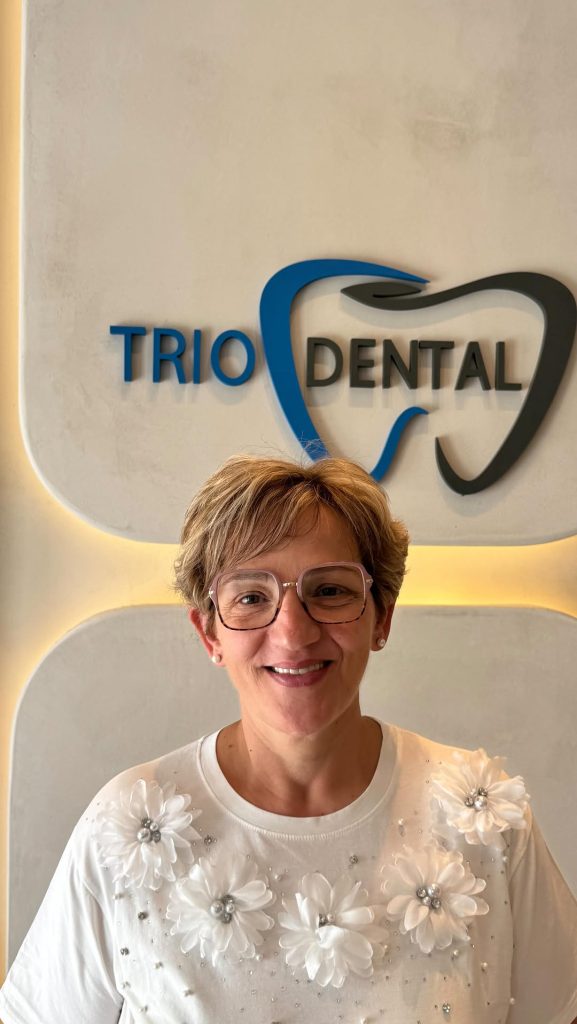 Donna sorridente con occhiali presso lo studio Trio Dental, con camicia bianca a fiori, in piedi accanto all'insegna del logo.