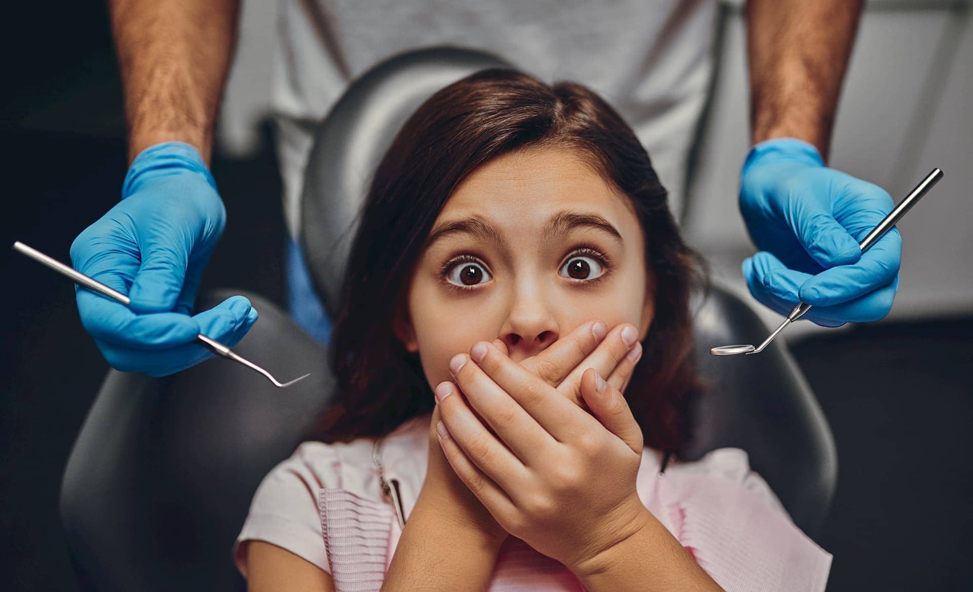 Bambino sulla poltrona del dentista con aria spaventata, che si copre la bocca, dentista che tiene gli strumenti con i guanti.