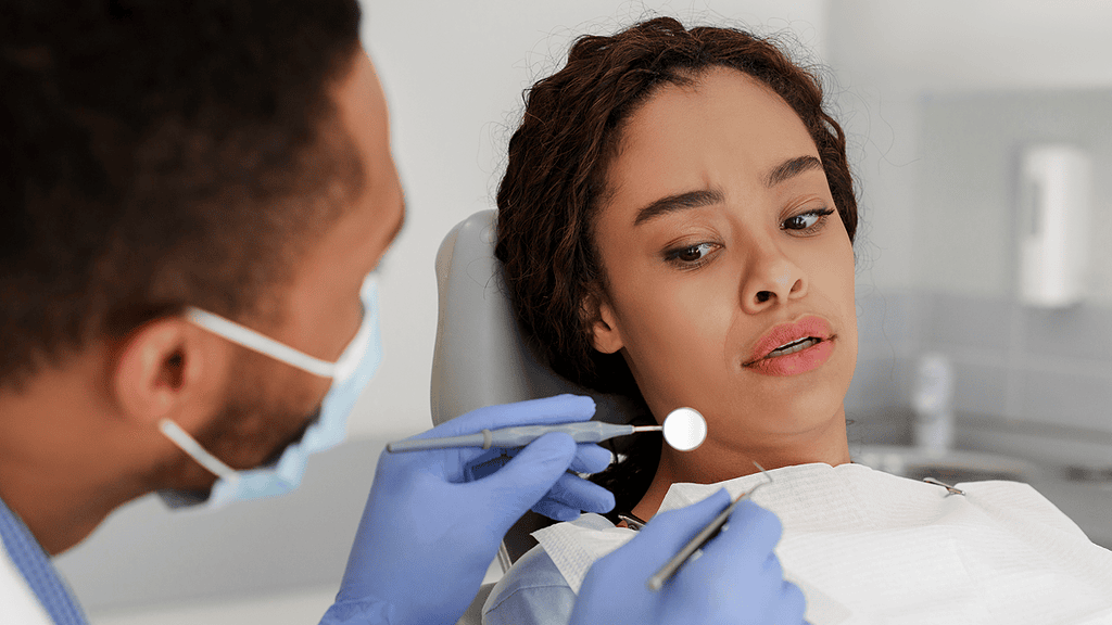 Patient at dentist's office looking concerned during dental exam, dentist holding dental tools, protective gear visible.