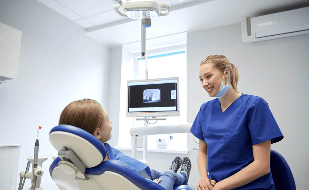 Dentista in camice blu sorride a giovane paziente sulla poltrona con radiografia sullo schermo.