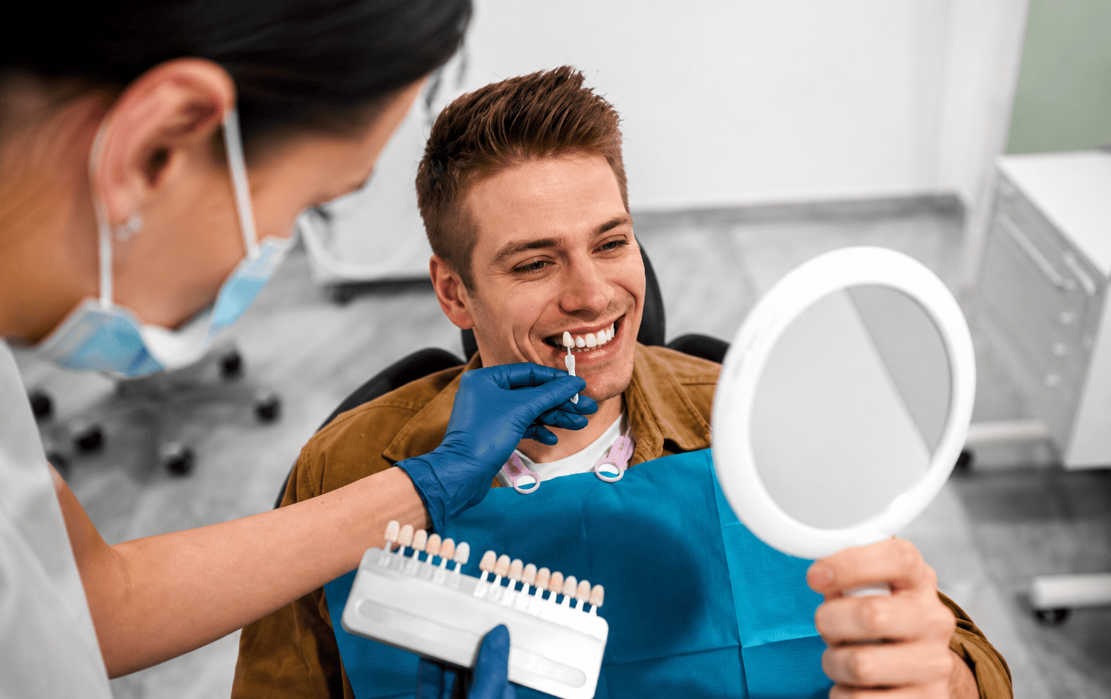 Paziente che sceglie il colore dei denti con il dentista, tenendo uno specchietto, sorridendo. Appuntamento dal dentista per sbiancamento dentale, consulto per bonding.