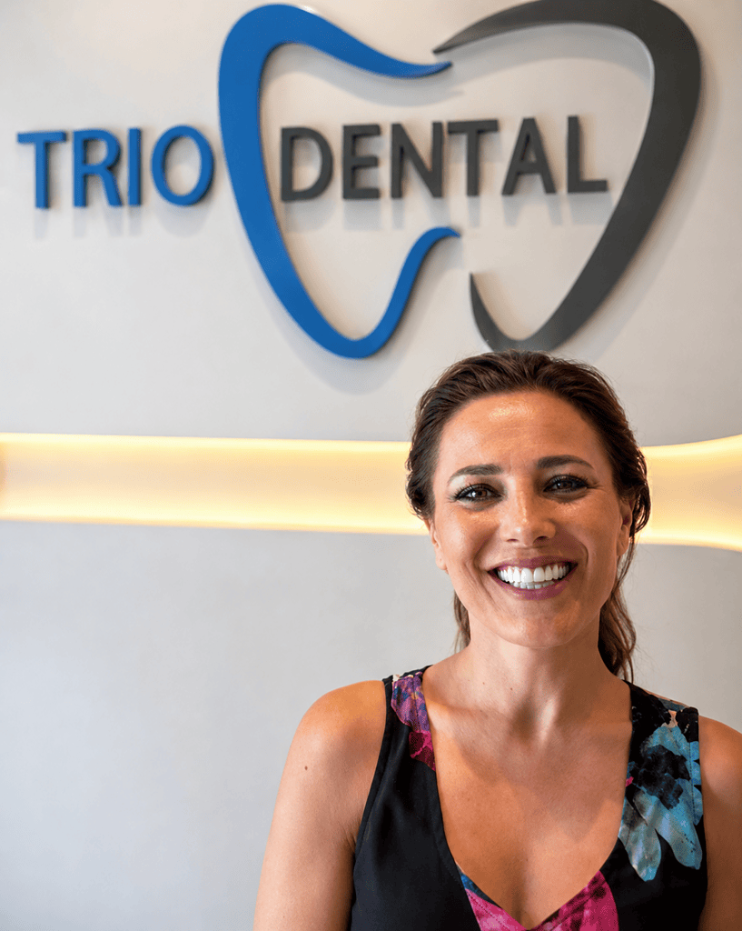 Smiling woman at Trio Dental clinic with logo in background, featuring tooth design on wall.