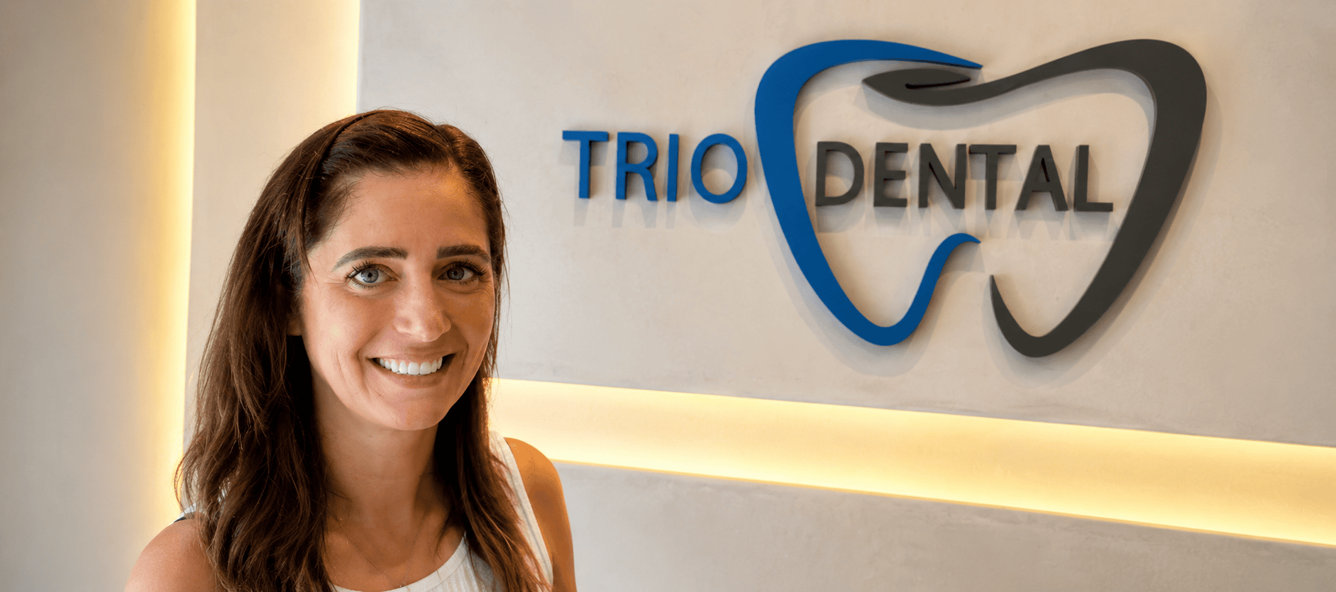 Smiling woman at Trio Dental office, logo on wall. Professional dental care.