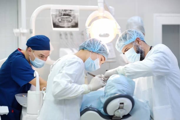 Dentists performing oral surgery in a dental clinic, wearing masks and surgical attire under bright lights.