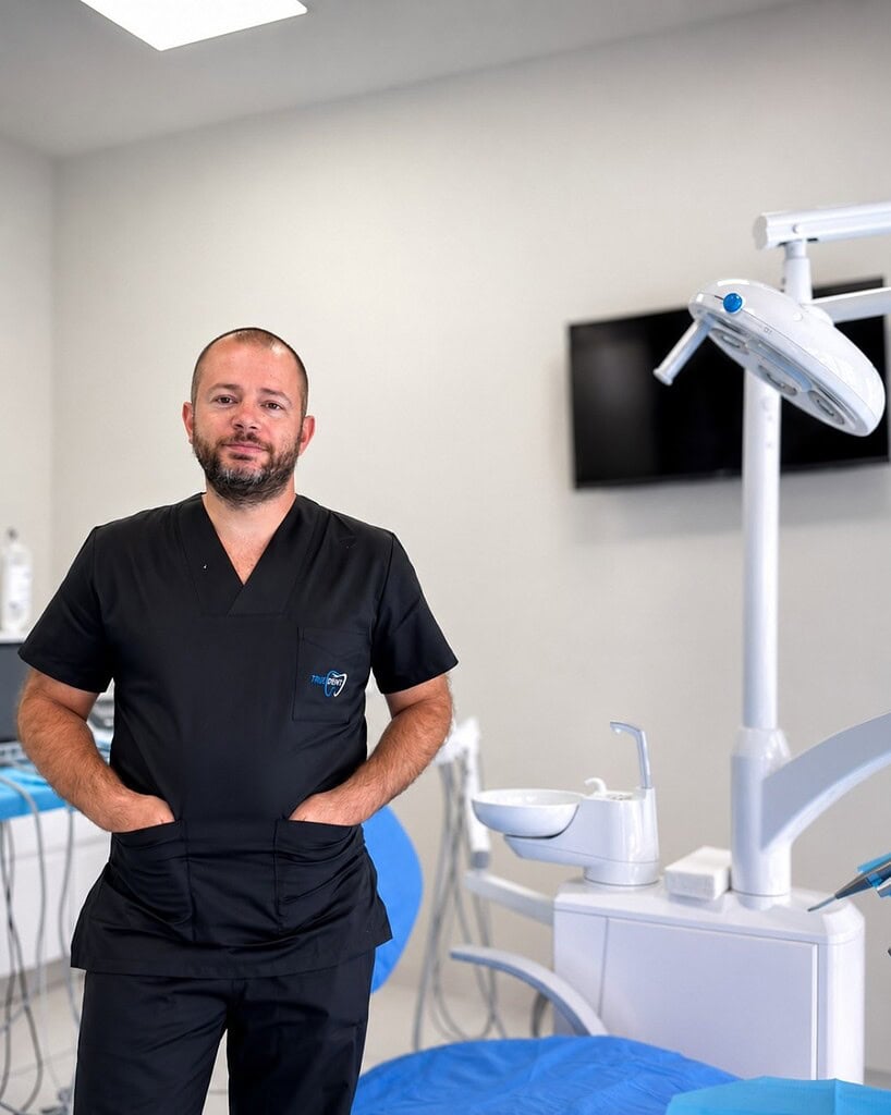 Dentiste en tenue médicale debout dans un cabinet dentaire moderne, à côté d’équipements dentaires et d’un fauteuil.