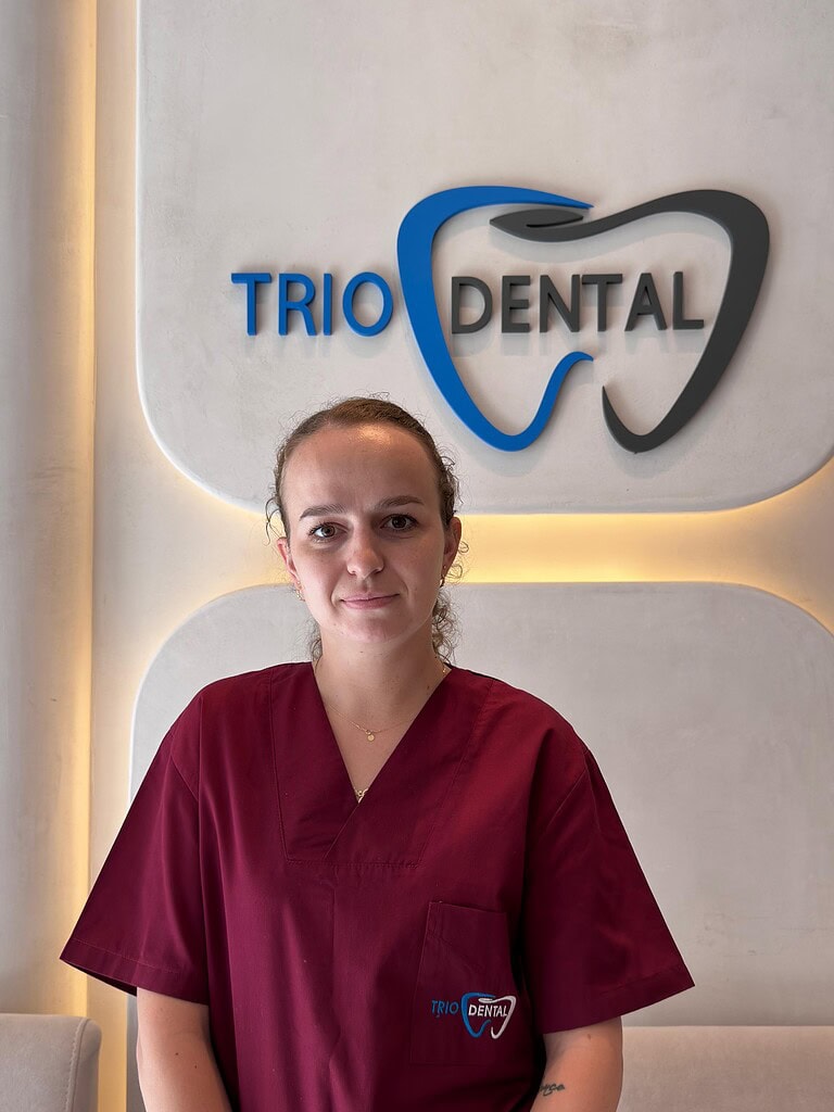 Dentist in maroon scrubs standing in front of Trio Dental logo on wall, showcasing modern dental clinic setting.