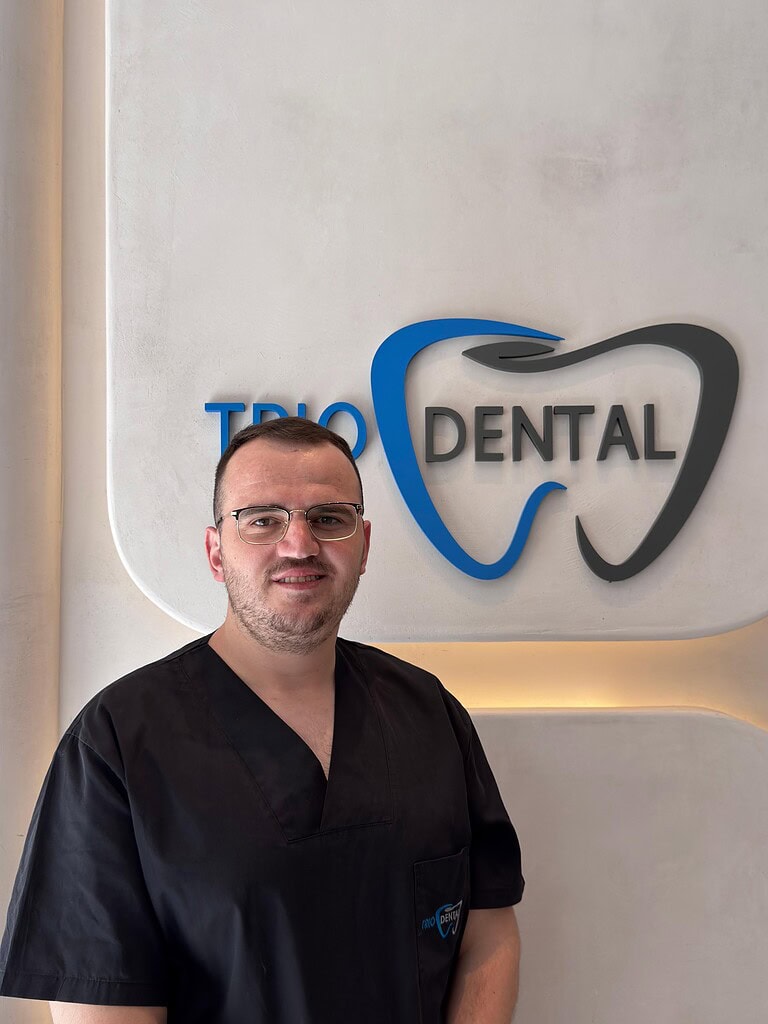 Dentist in scrubs standing in front of Trio Dental logo on clinic wall.