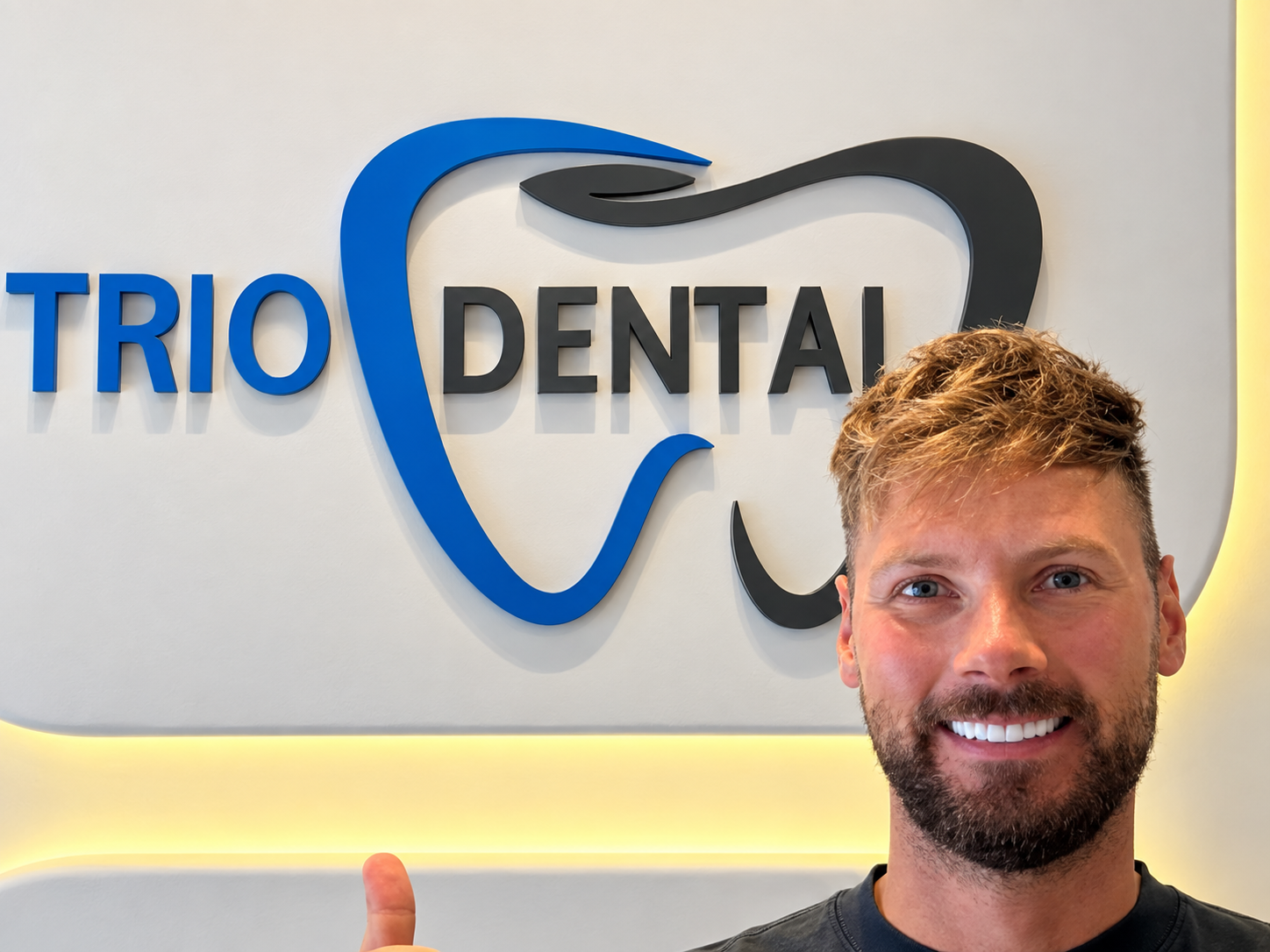 Smiling man in front of Trio Dental logo, giving a thumbs up, at a dental clinic.