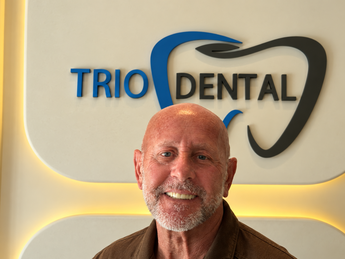 Man smiling in front of Trio Dental logo, featuring a modern design with tooth imagery.